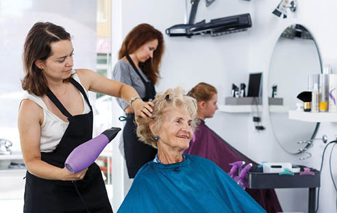 Professional female stylist making hairdo for senior woman in salon, using hair dryer 