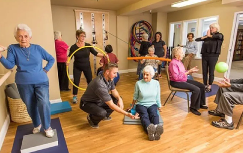 A group of people are in a room, some of them are sitting on chairs, and others are standing. A man is kneeling down next to a woman who is sitting on the floor. The group appears to be engaged in an exercise or physical activity. AI generated content