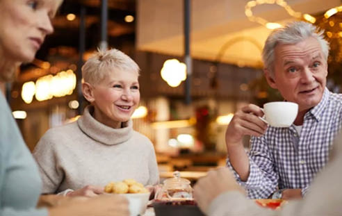 Three people are sitting at a table, enjoying a meal together. They are smiling and having a good time. A man is holding a cup of coffee, and there are several donuts on the table. The scene appears to be a casual gathering of friends or family. AI generated content